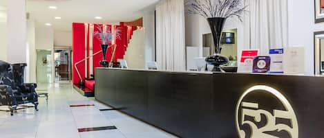 Un espace d’accueil d’hôtel au design moderne, avec un comptoir noir orné du logo de l’établissement, un mur d’accent rouge et un escalier menant à un autre étage.