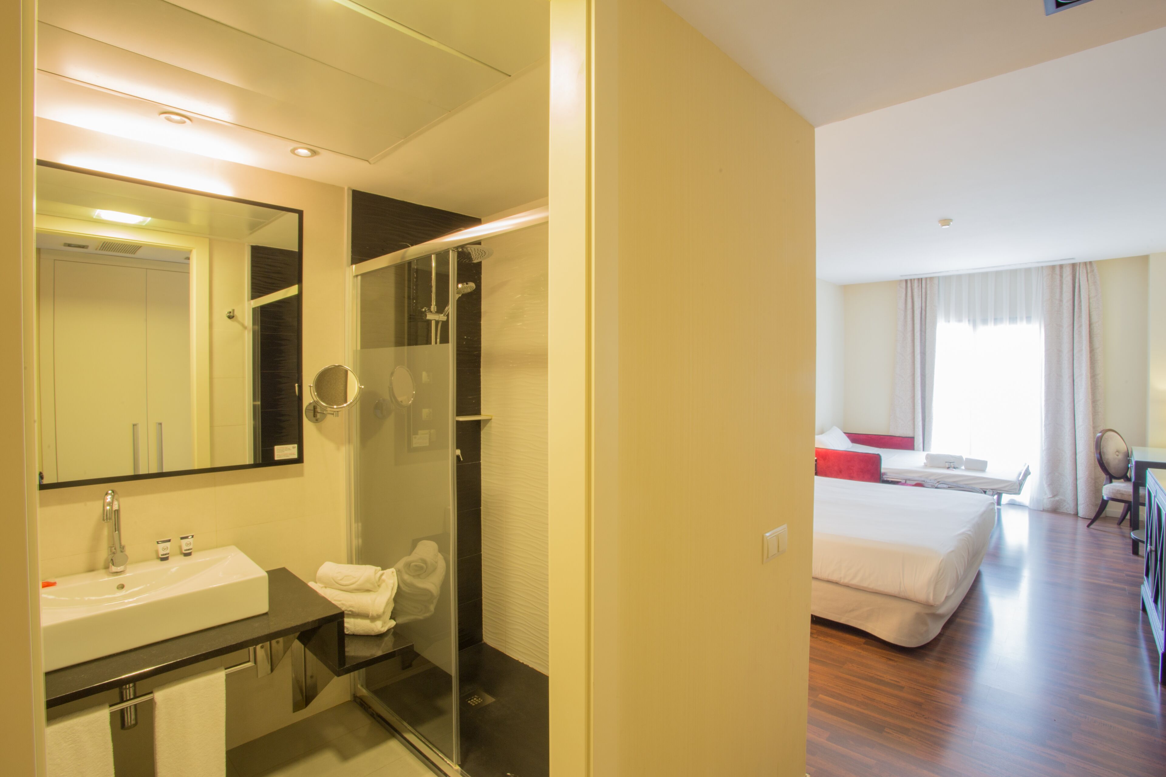 Quarto de hotel com banheiro, apresentando pia, espelho e chuveiro. O quarto possui uma cama e uma escrivaninha com cadeira.