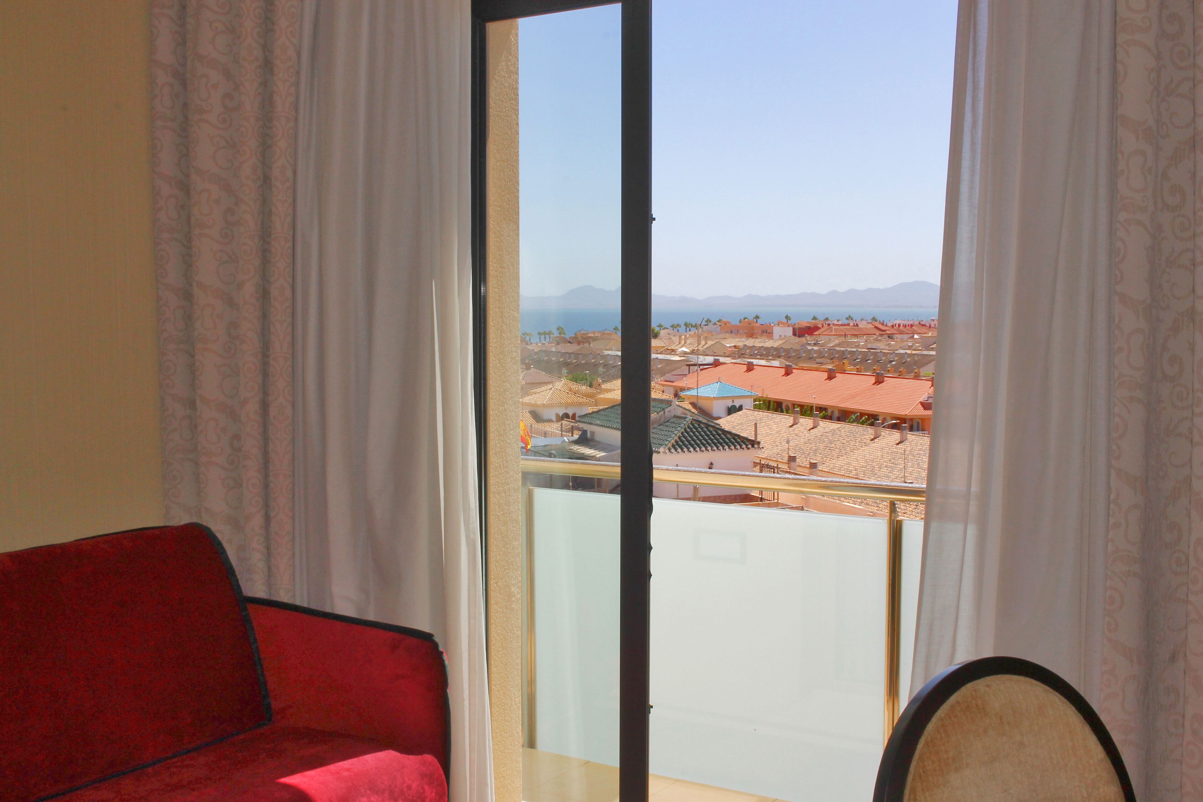 Ein Zimmer mit einem roten Sofa, einem großen Fenster mit weißen Vorhängen und Blick auf eine Küstenlandschaft mit Gebäuden und Bergen.