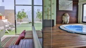 Una sala de estar moderna con un ventanal que da a un patio, piso de madera, una bañera de hidromasaje redonda y una escultura montada en la pared.