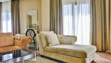 Una sala de estar con un sofá beige, una mesa de centro negra y una mesa redonda con una silla. Un espejo cuelga en la pared, y la habitación tiene ventanas grandes con cortinas.