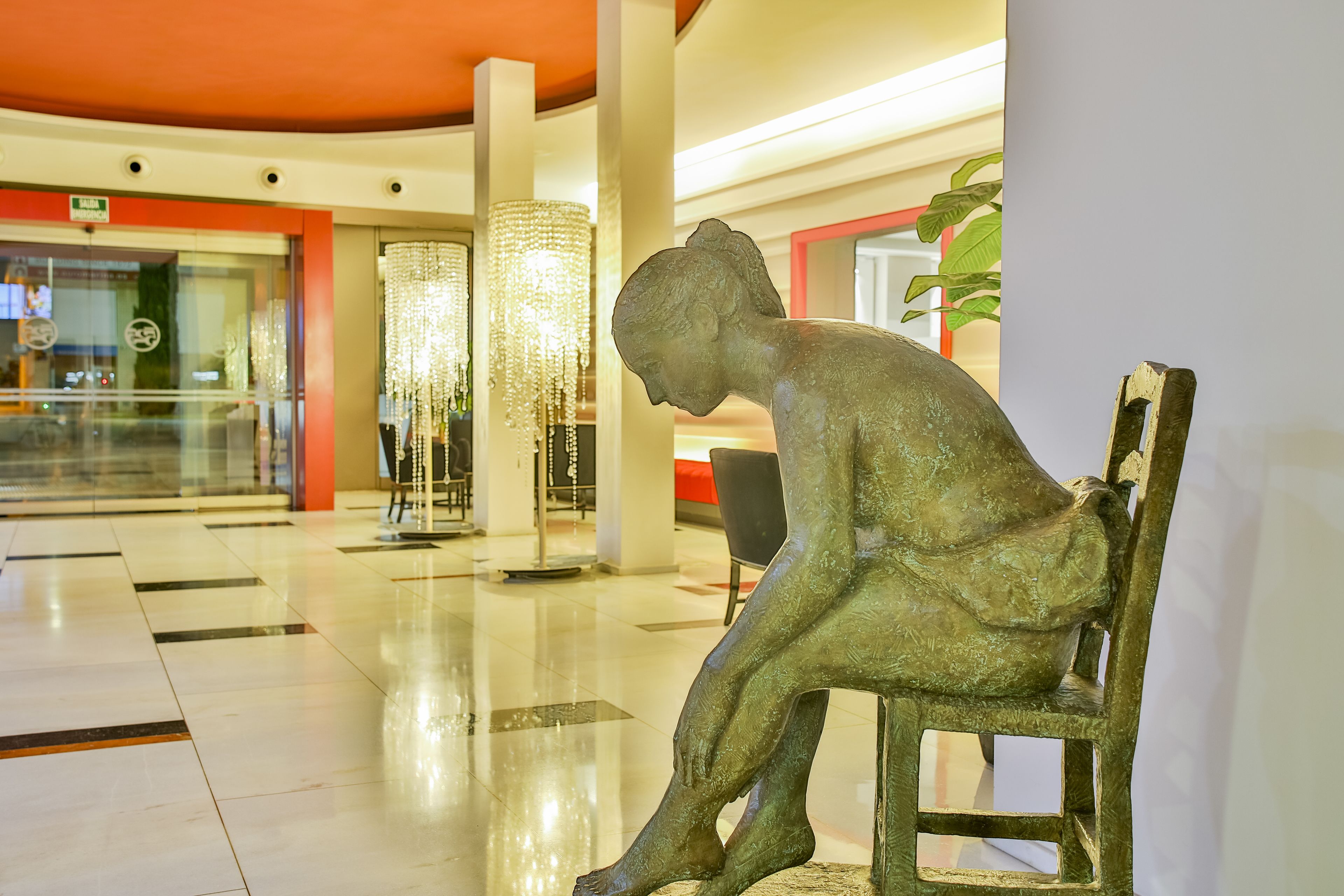 Una estatua de una figura sentada en un espacio interior público con suelo pulido, una araña y una planta de fondo.