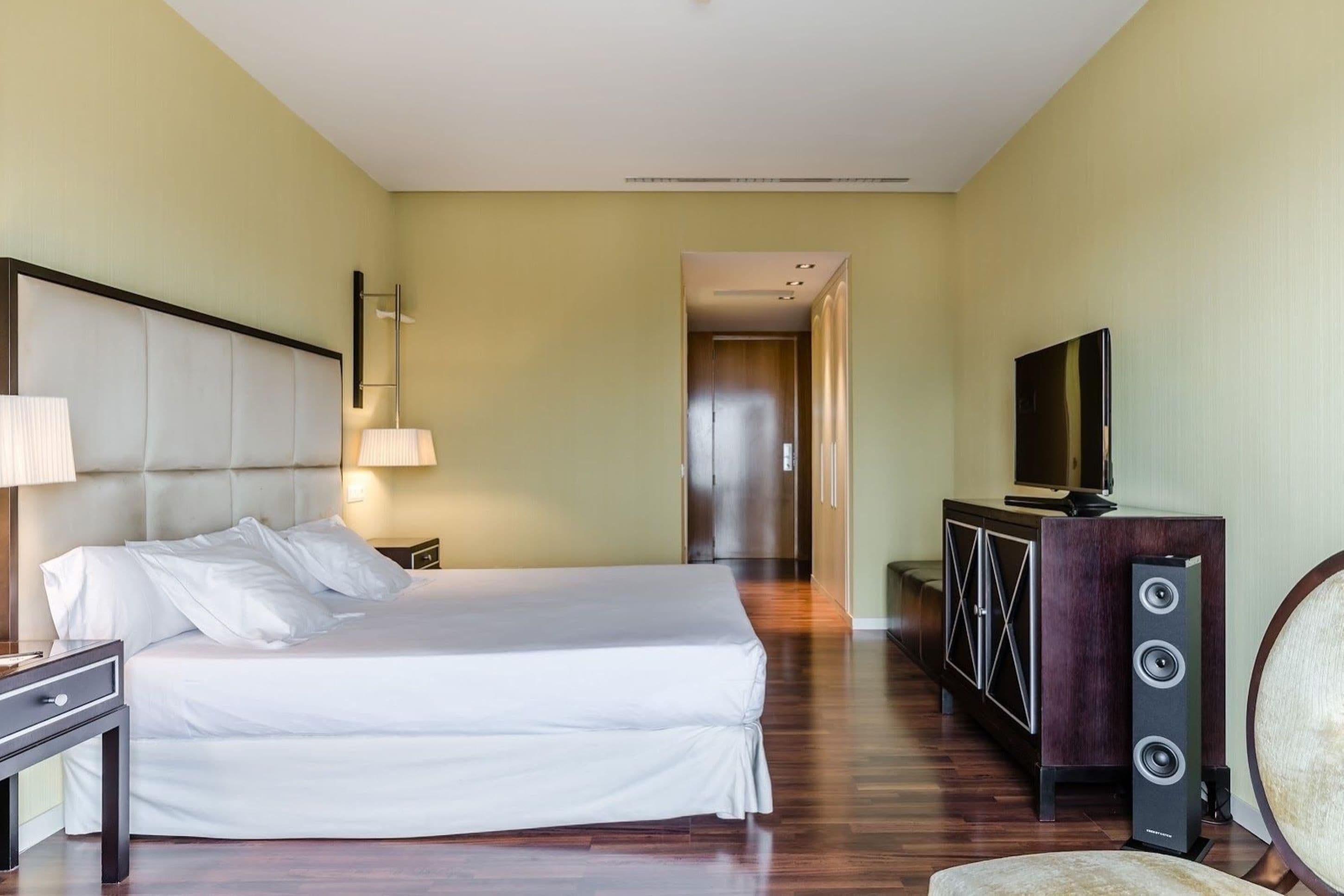 A hotel room with a large bed, a TV, a cabinet with speakers, and a hallway leading to another room.