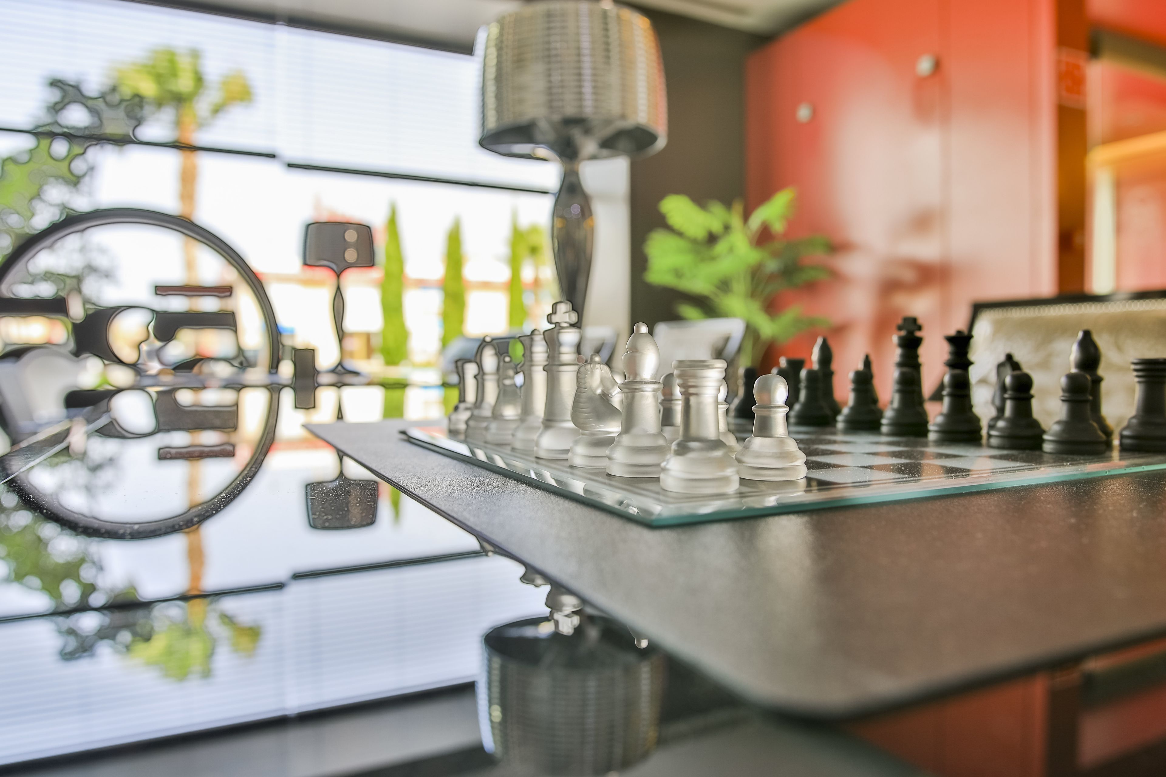 A chessboard on a reflective surface with a lamp and a plant in the background.