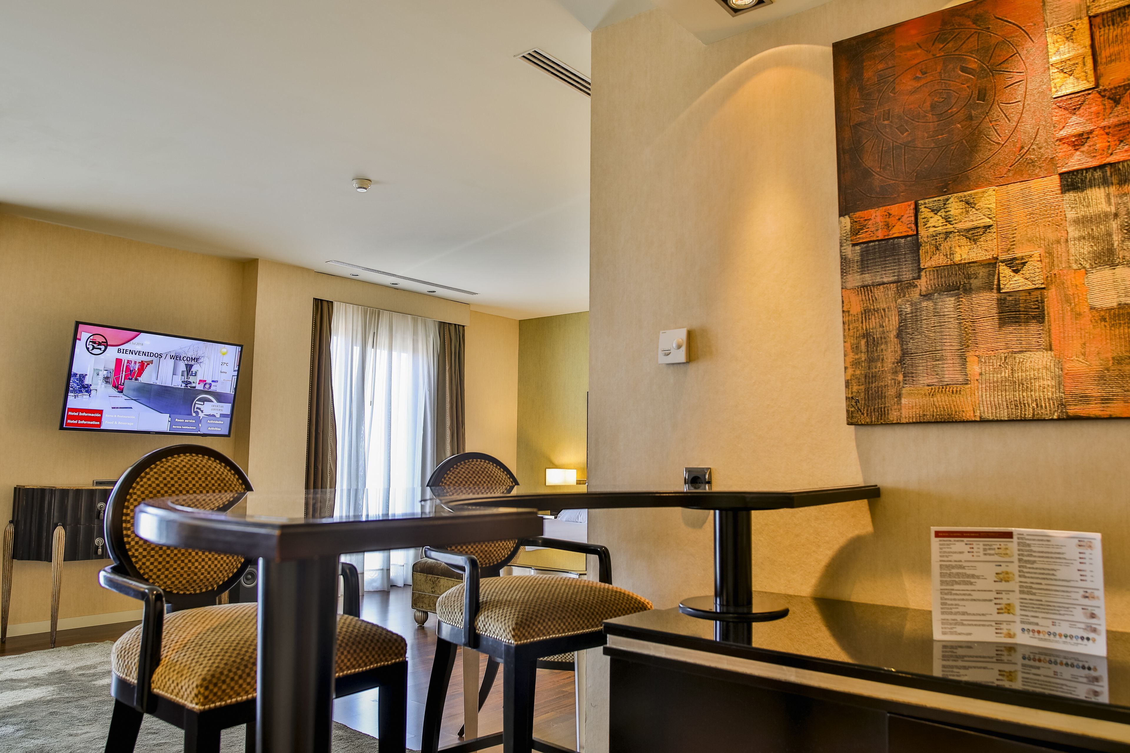 Una habitación de hotel con zona de comedor, un televisor de pantalla plana mostrando un mensaje de bienvenida y una obra de arte abstracta colgada en la pared.