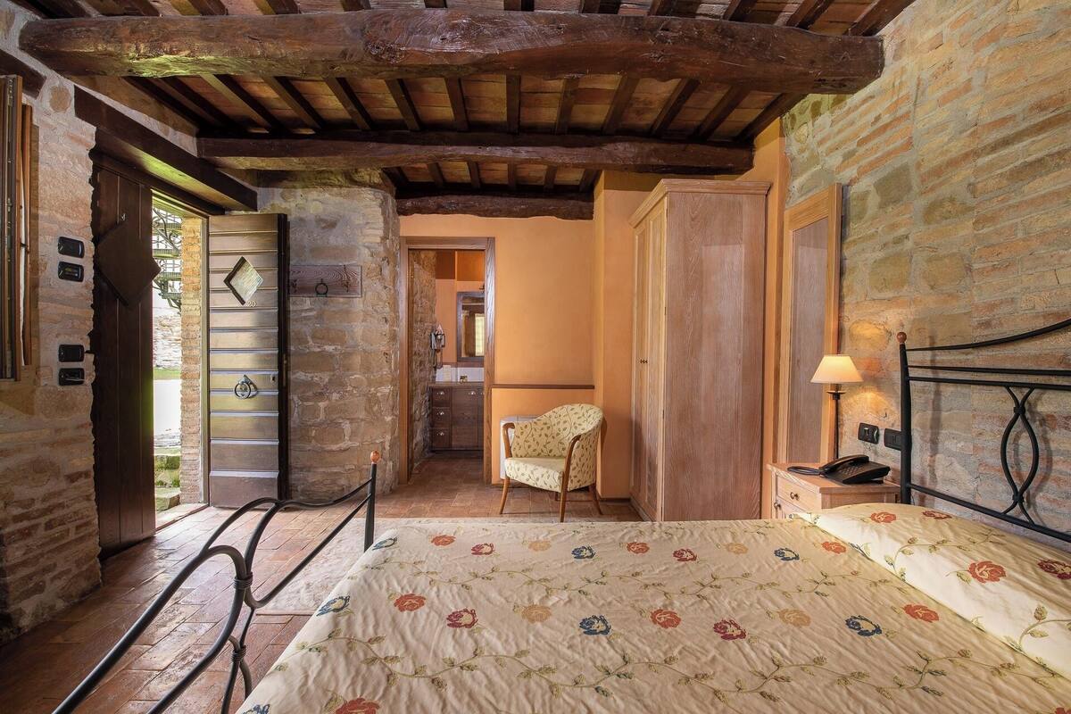 Een slaapkamer met een houten plafond, stenen muren, een bed met een bloemenbedekking, een stoel en een nachtkastje met een lamp.
