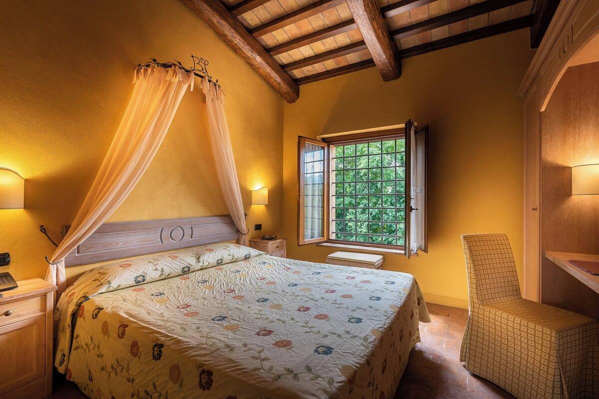 Een slaapkamer met hemelbed, een raam met uitzicht op groen, nachtkastje en een stoel.