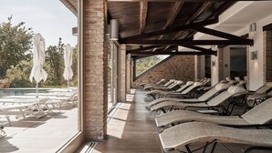 A sunlit outdoor lounge area with wicker lounge chairs and umbrellas, a pool, and a stone wall.