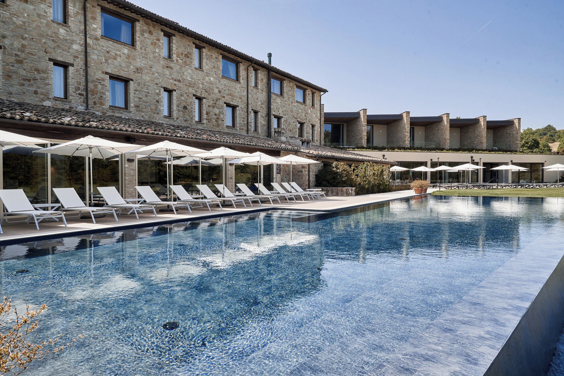 A pool with clear water, surrounded by white lounge chairs and umbrellas, and a stone building in the background.