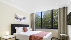 Ein Hotelzimmer mit einem großen Bett, einem Nachttisch, einem Fenster mit Blick auf Bäume und einem Bild an der Wand.