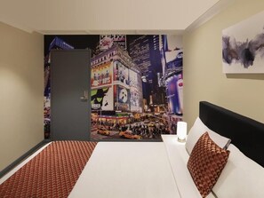 A hotel room with a bed, a nightstand, a lamp, a painting, and a wall mural of a cityscape. - BreakFree on Broadway Sydney (Glebe)