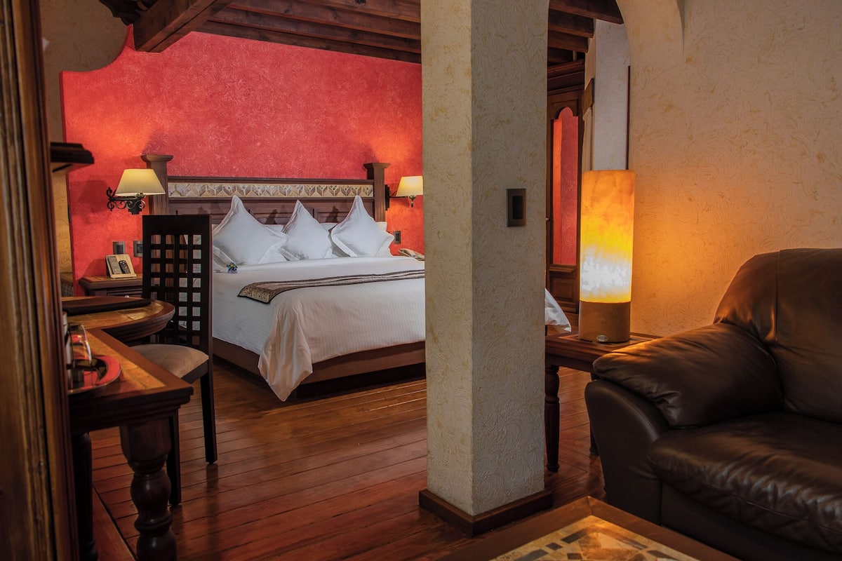 A hotel room with a bed, two bedside lamps, a wooden nightstand, a leather armchair, and a wooden floor.
