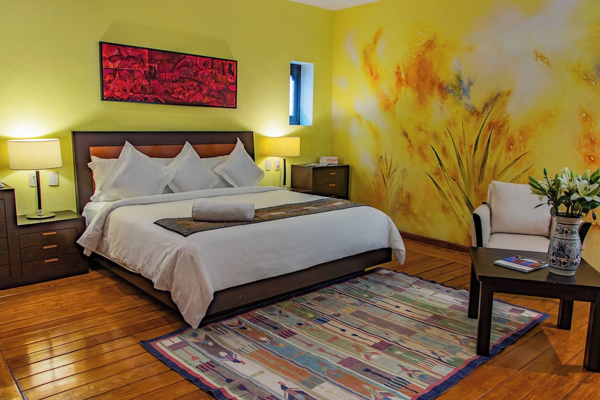 A neatly made bed with white linens and pillows, a wooden floor, a colorful rug, a nightstand with a lamp, a chair with a vase of flowers, and a painting on the wall.