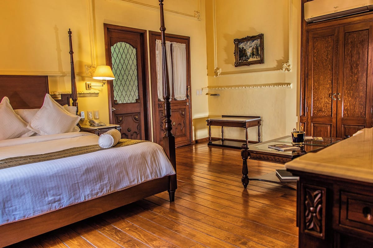 A hotel room with a four-poster bed, wooden flooring, a small table with a lamp, a painting on the wall, and a large wardrobe.
