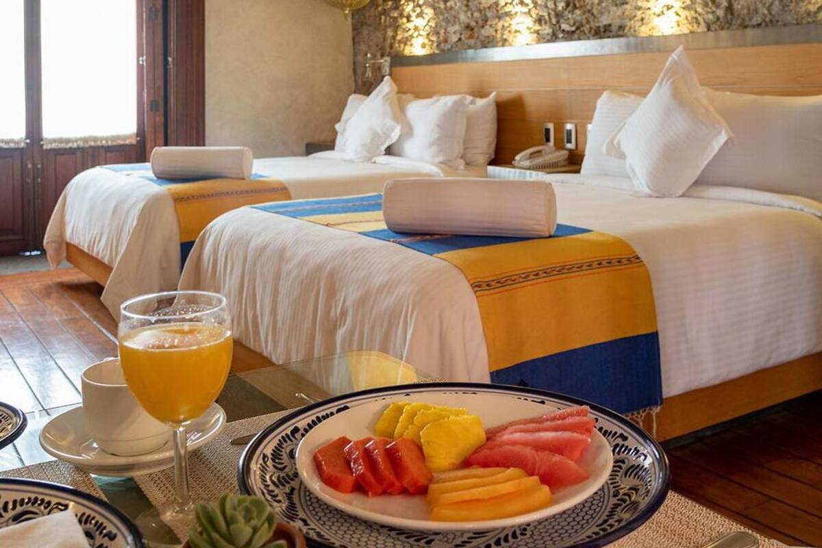 A hotel room with two beds, a breakfast table with fruit and a glass of orange juice, and a decorative wall.