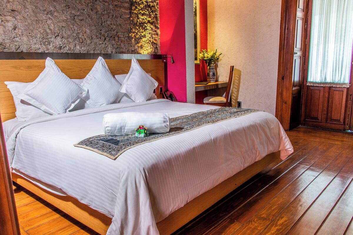 A neatly made bed with white linens and pillows, a bedside table with a vase and a small figurine, and a wooden floor.