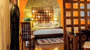 Un dormitorio con pared de piedra, cabecera de madera, cama con ropa blanca, una silla de madera y una mesita con una lámpara.