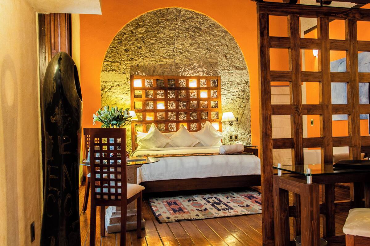 A bedroom with a stone wall, a wooden headboard, a bed with white linens, a wooden chair, and a small table with a lamp.