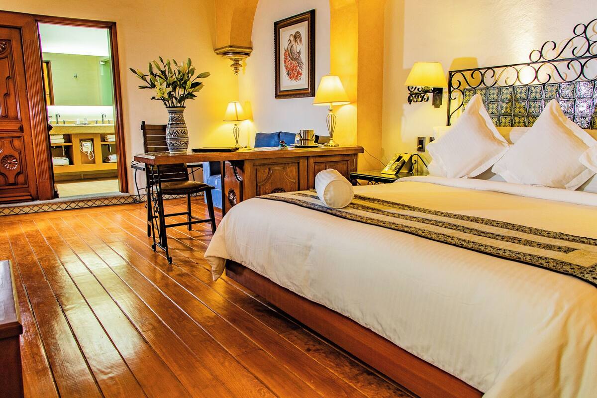 A hotel room with a wooden floor, a bed with white linens, a desk with two lamps, and a view of the bathroom through an open door.