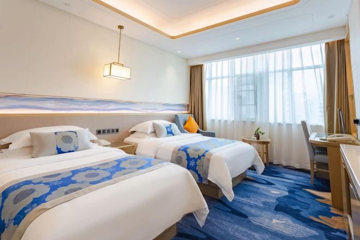 Habitación de hotel con dos camas, un escritorio, una silla, una lámpara y un ventanal grande con cortinas.