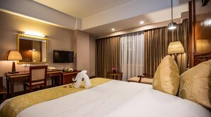 A hotel room with a bed, a desk, a television, a mirror, a window with curtains, and a lamp.