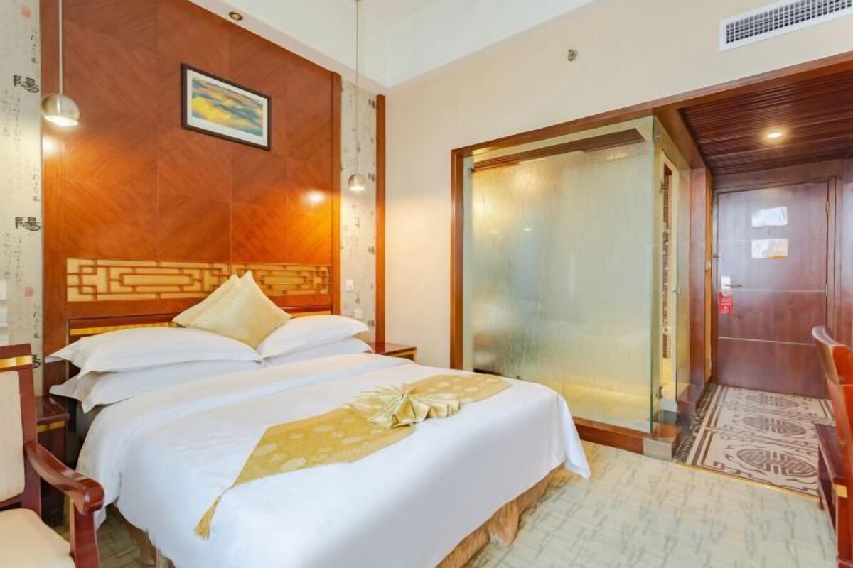 A hotel room with a large bed, a wooden headboard, a glass door, and a small table with a chair.