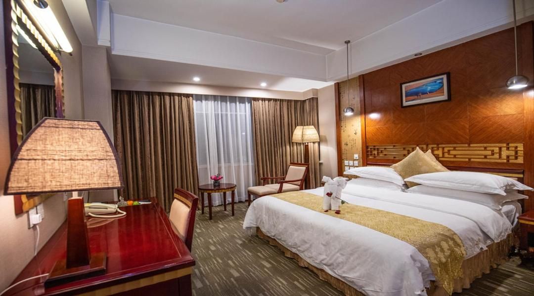 A hotel room with a bed, a desk, a chair, a lamp, and a painting on the wall.