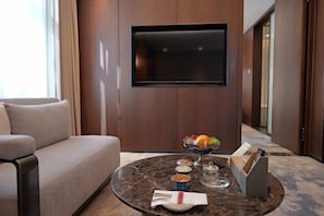 Een moderne hotelkamer met een bank, een ronde marmeren salontafel met snacks, een aan de muur bevestigde televisie en een gang die naar een andere kamer leidt.