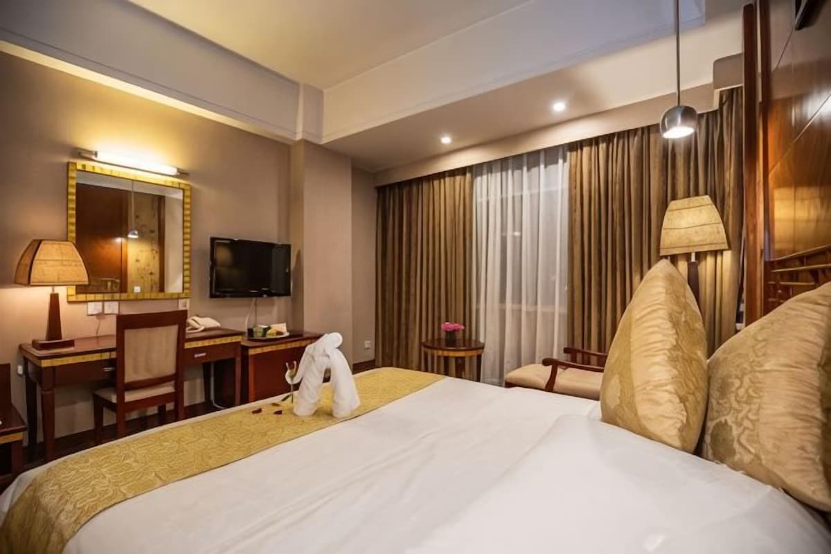 Habitación de hotel con cama, escritorio con teléfono, televisor, espejo, mesita de noche con lámpara y ventana con cortinas.