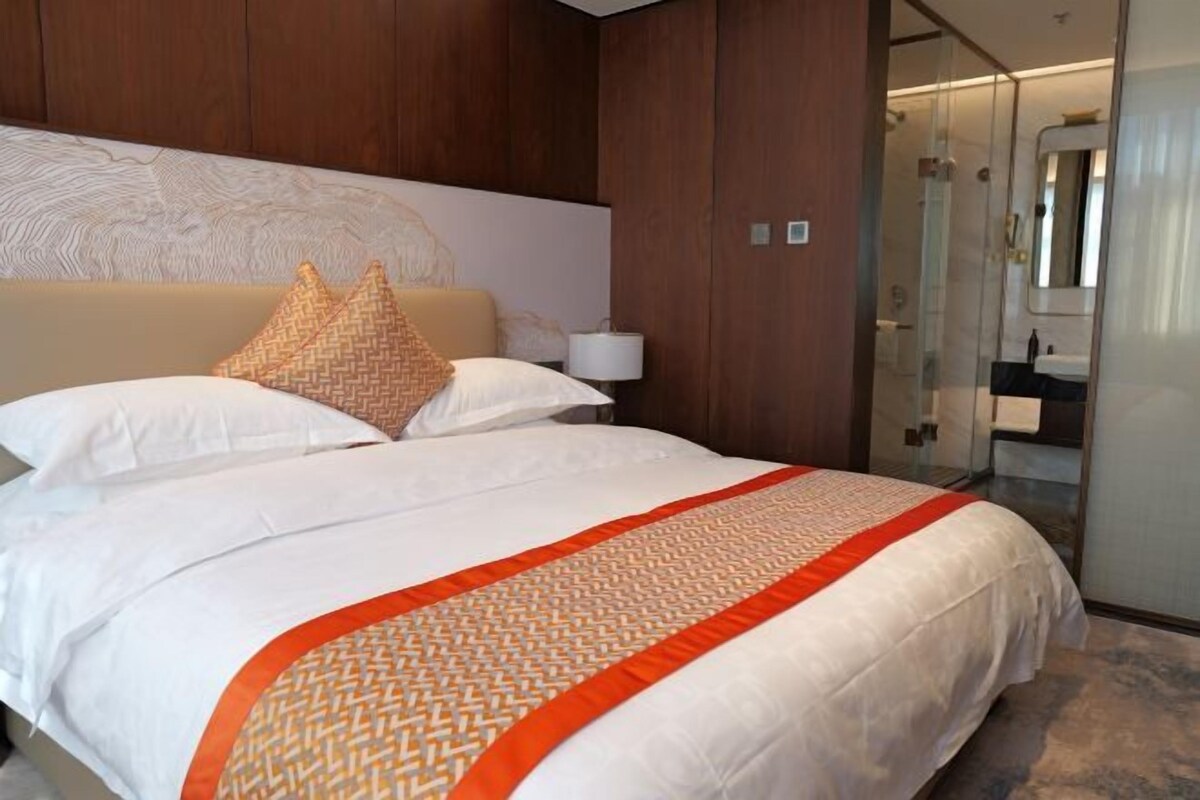 Una habitación de hotel con una cama grande, un cabecero con diseño texturizado, una mesita de noche con lámpara y un box de ducha de vidrio.