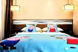 Habitación de hotel con una cama, almohadones con personajes de caricatura, una colcha azul con personajes similares y un cabecero moderno.