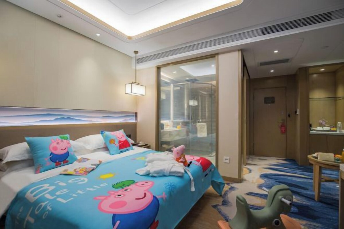 A hotel room with a bed featuring cartoon character bedding, a desk with a chair, and a bathroom with a glass shower enclosure.