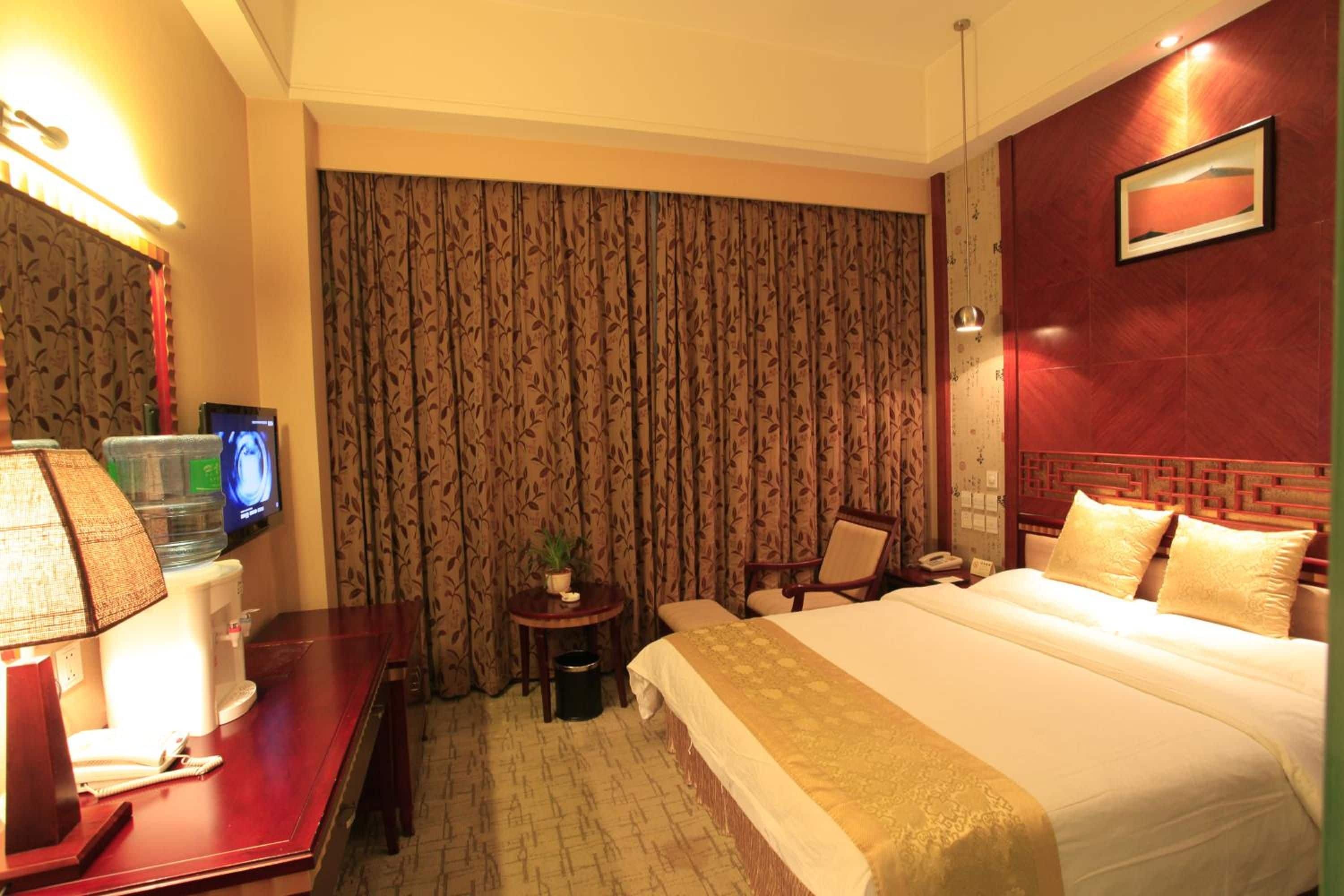 A hotel room with a bed, a desk with a computer, a chair, a small table with a plant, and a wall-mounted TV.