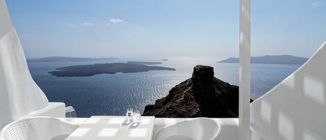 A white outdoor seating area with a table and chairs, overlooking a scenic view of the sea and islands.