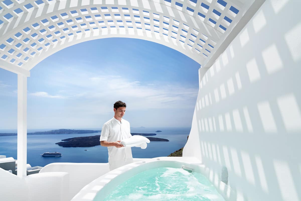 A man in a white shirt standing by a white infinity pool with a clear view of the sea and islands in the background.