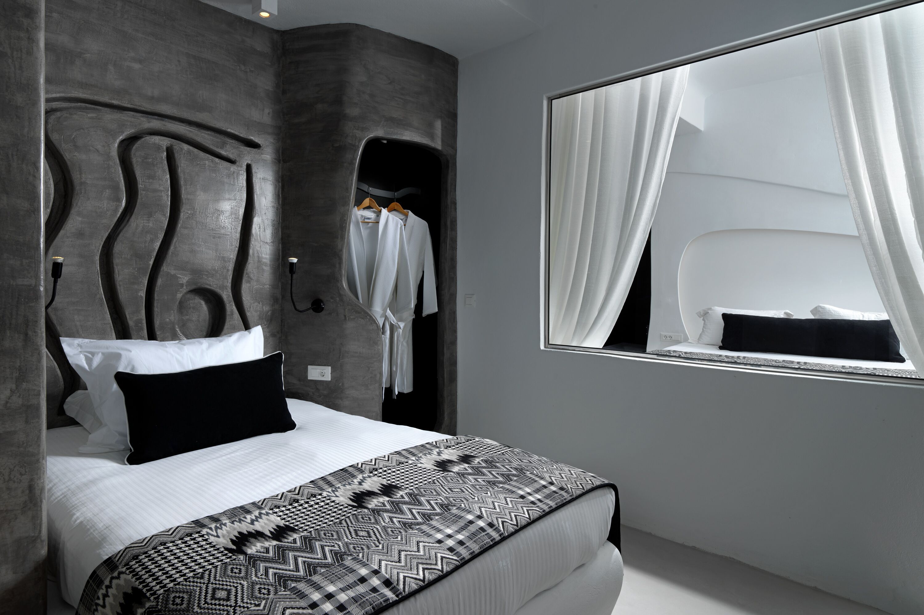 A modern bedroom with a large bed, a headboard with a unique design, a window with sheer curtains, and a wardrobe with hanging clothes.