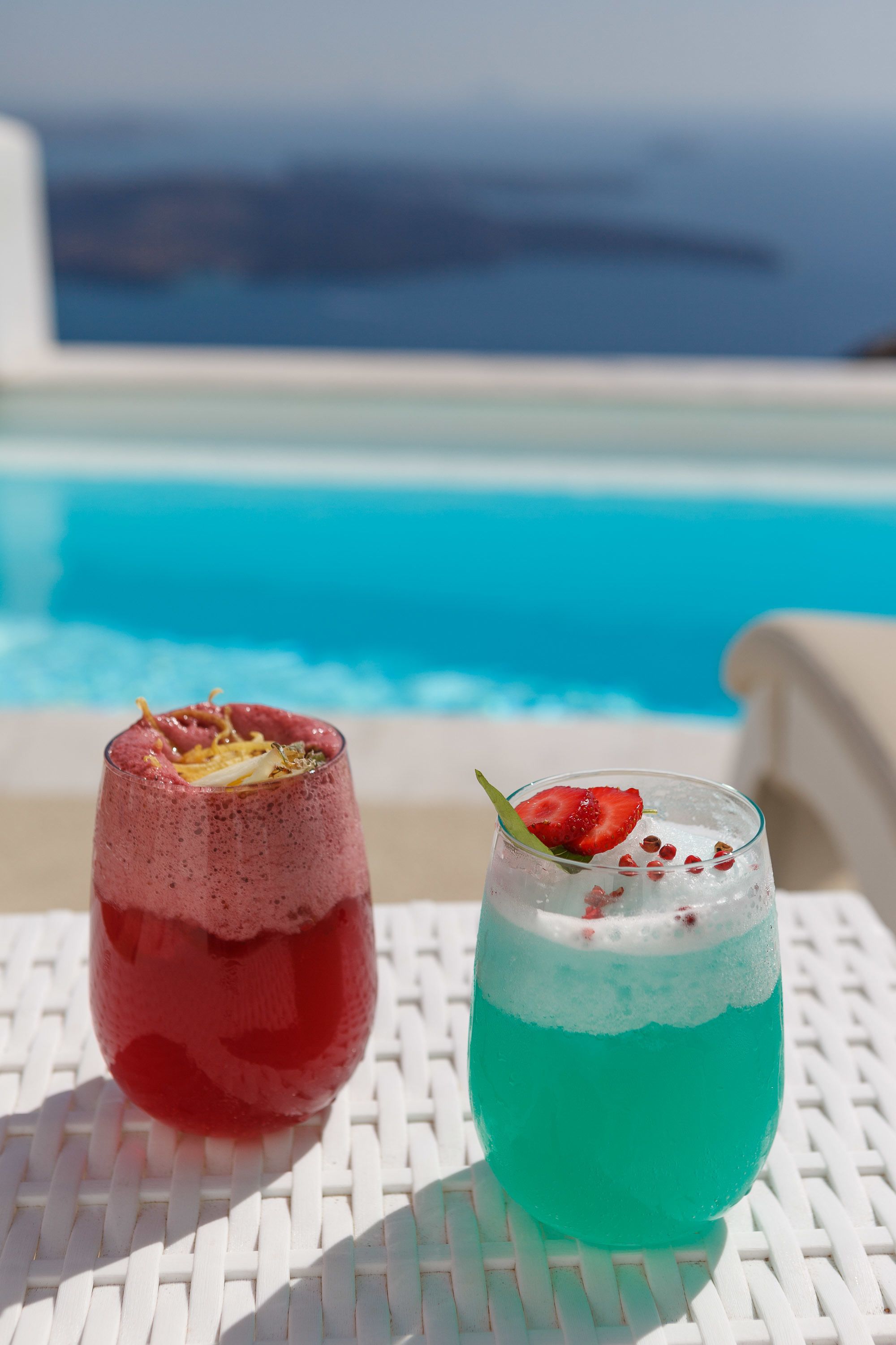 Two drinks on a white table by a pool with a clear view of the ocean.