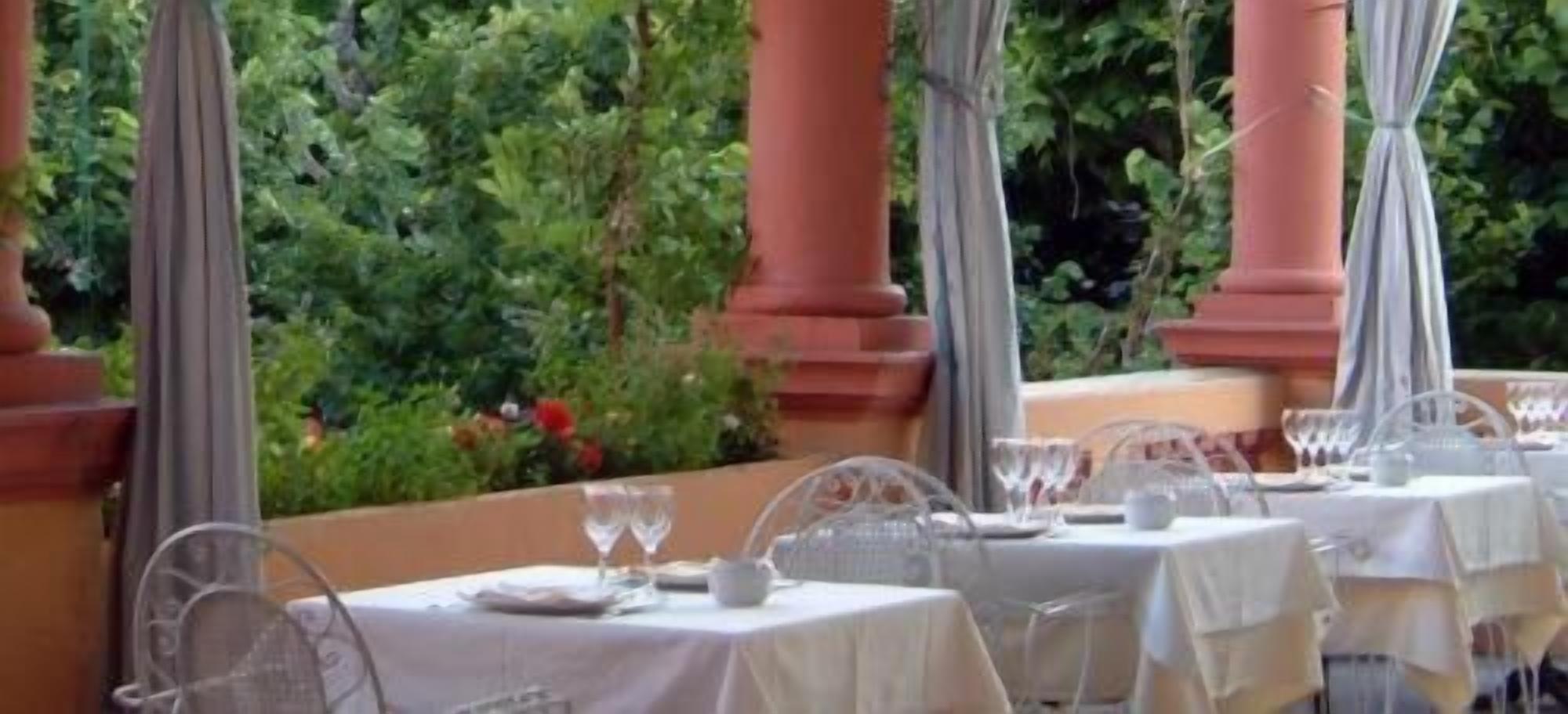 An outdoor dining area with tables set for two, surrounded by greenery and columns.
