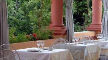 An outdoor dining area with tables set for two, surrounded by greenery and columns.