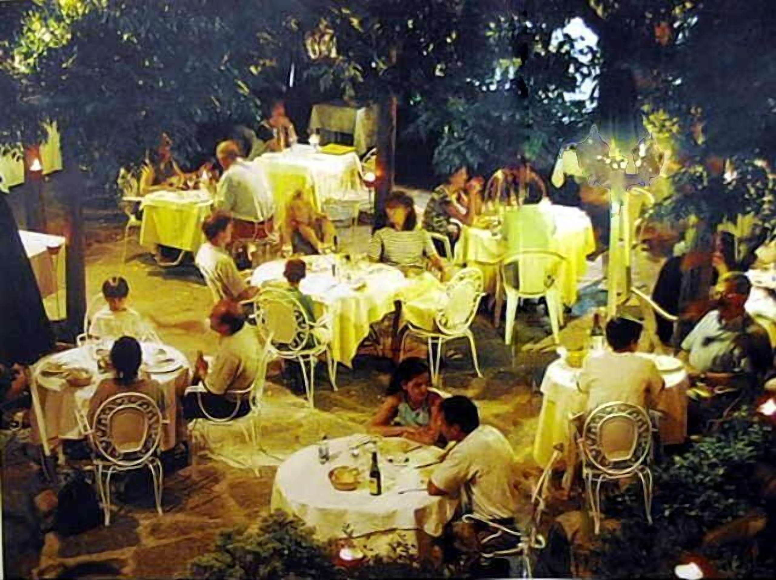 Un área de comedor al aire libre con mesas y sillas redondas, rodeada de árboles y cubierta con manteles amarillos.