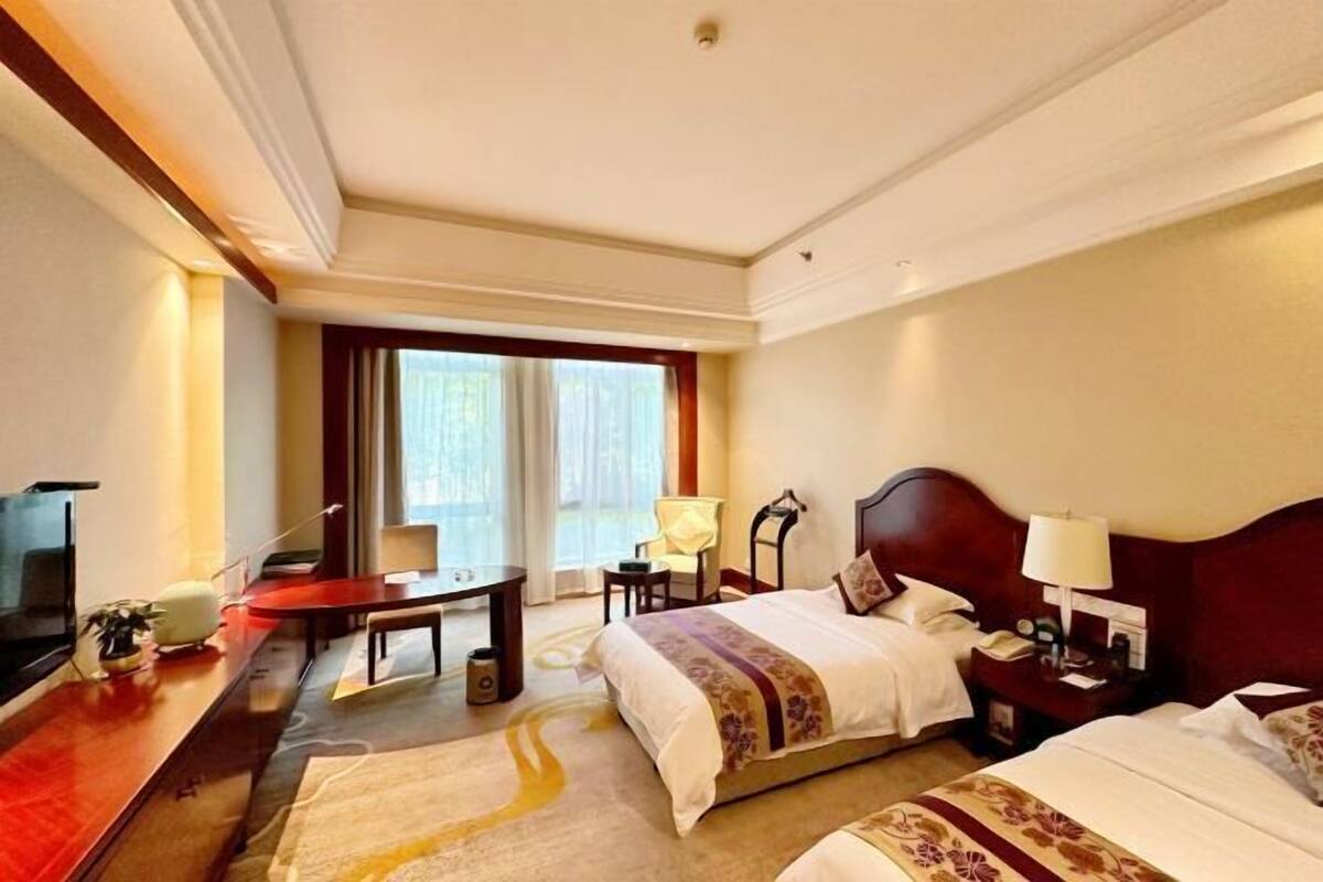 A hotel room with a bed, bedside tables, a desk, a chair, a TV, and a large window.