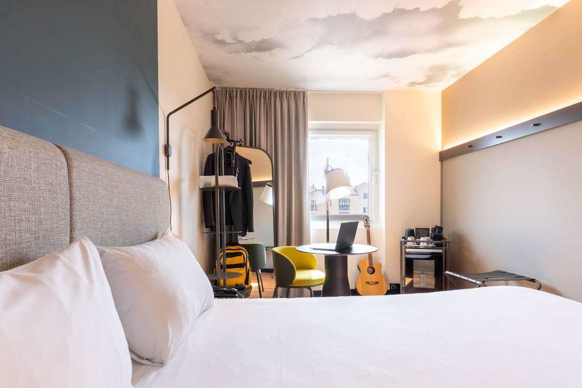 Une chambre d’hôtel avec un lit, un bureau avec un ordinateur portable, une guitare, une chaise et une fenêtre avec des rideaux.