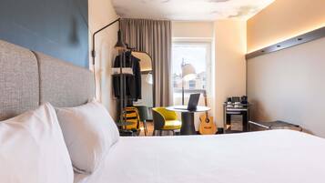 A hotel room with a bed, a desk with a laptop, a guitar, a chair, and a window with curtains.