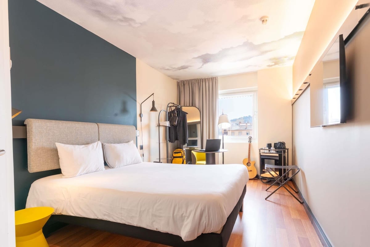 Une chambre d’hôtel avec un lit, un bureau, une chaise, une guitare et une fenêtre donnant sur un beau paysage.