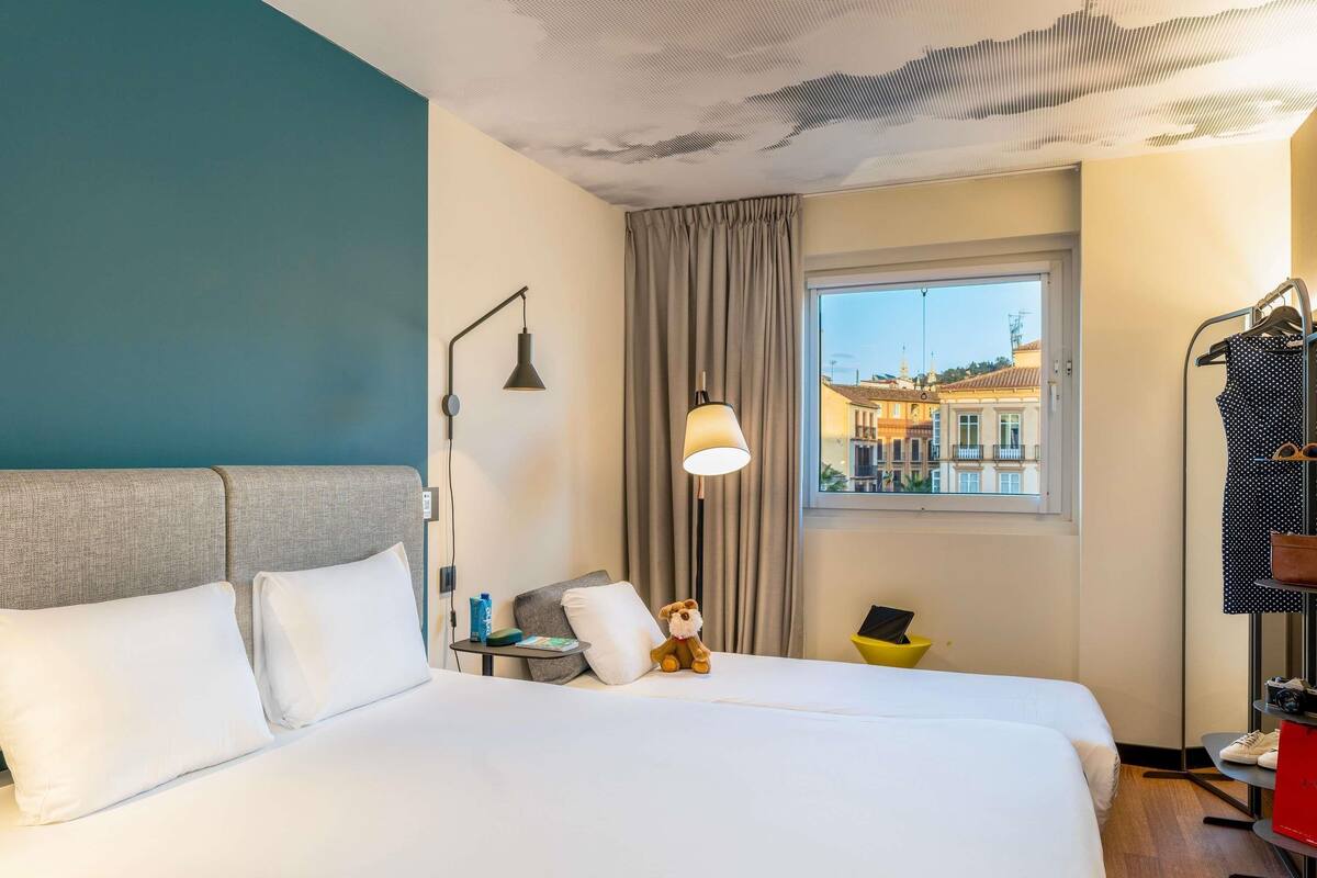 Une chambre d’hôtel avec un grand lit, une table de chevet, un ours en peluche, une fenêtre donnant sur des immeubles et un portant à vêtements avec un sac rouge.