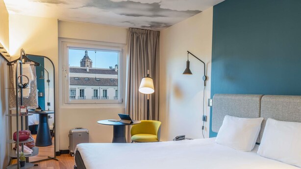 Een hotelkamer met een bed, een stoel, een tafel, een lamp, een koffer en uitzicht op een gebouw.