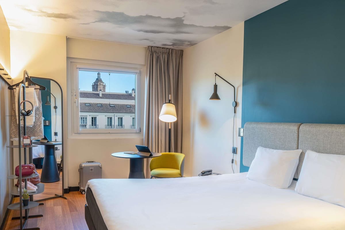Une chambre d’hôtel avec un lit, une chaise, une table, une lampe, une valise et une vue sur un bâtiment à travers la fenêtre.