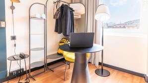 Een moderne hotelkamer met een laptop op een tafel, een staande plank met kleding en een tas, een telefoon op een bijzettafeltje, en een uitzicht op de stad door het raam.