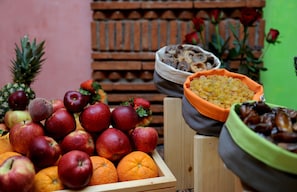 Una caixa de fusta plena de pomes vermelles i taronges, un bol de fruits secs i una pinya.