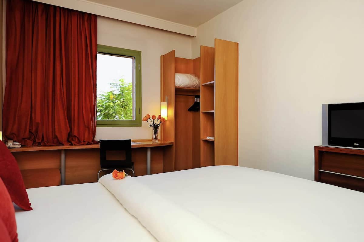 A hotel room with a bed, a TV, a desk with a chair, a wardrobe, and a window with curtains.